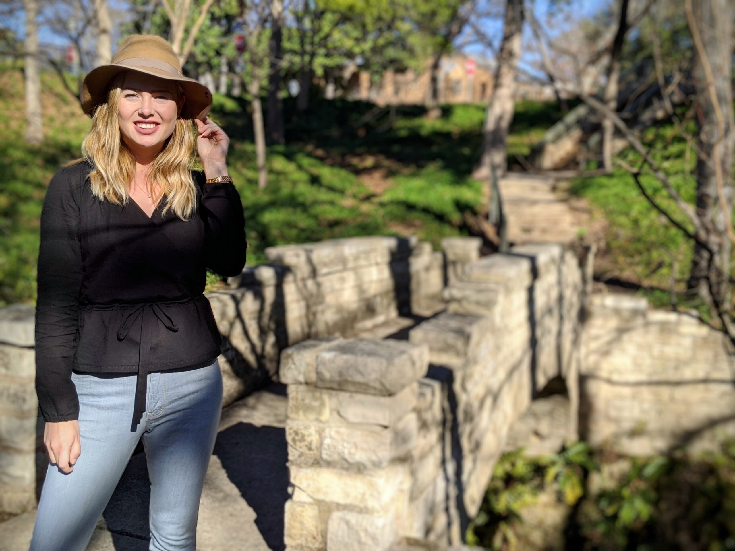 Elizabeth stands near a stone bridge in a park in the wintertime. She is wearing a tan felt hat, and a handmade black wrap top and light colored denim jeans.