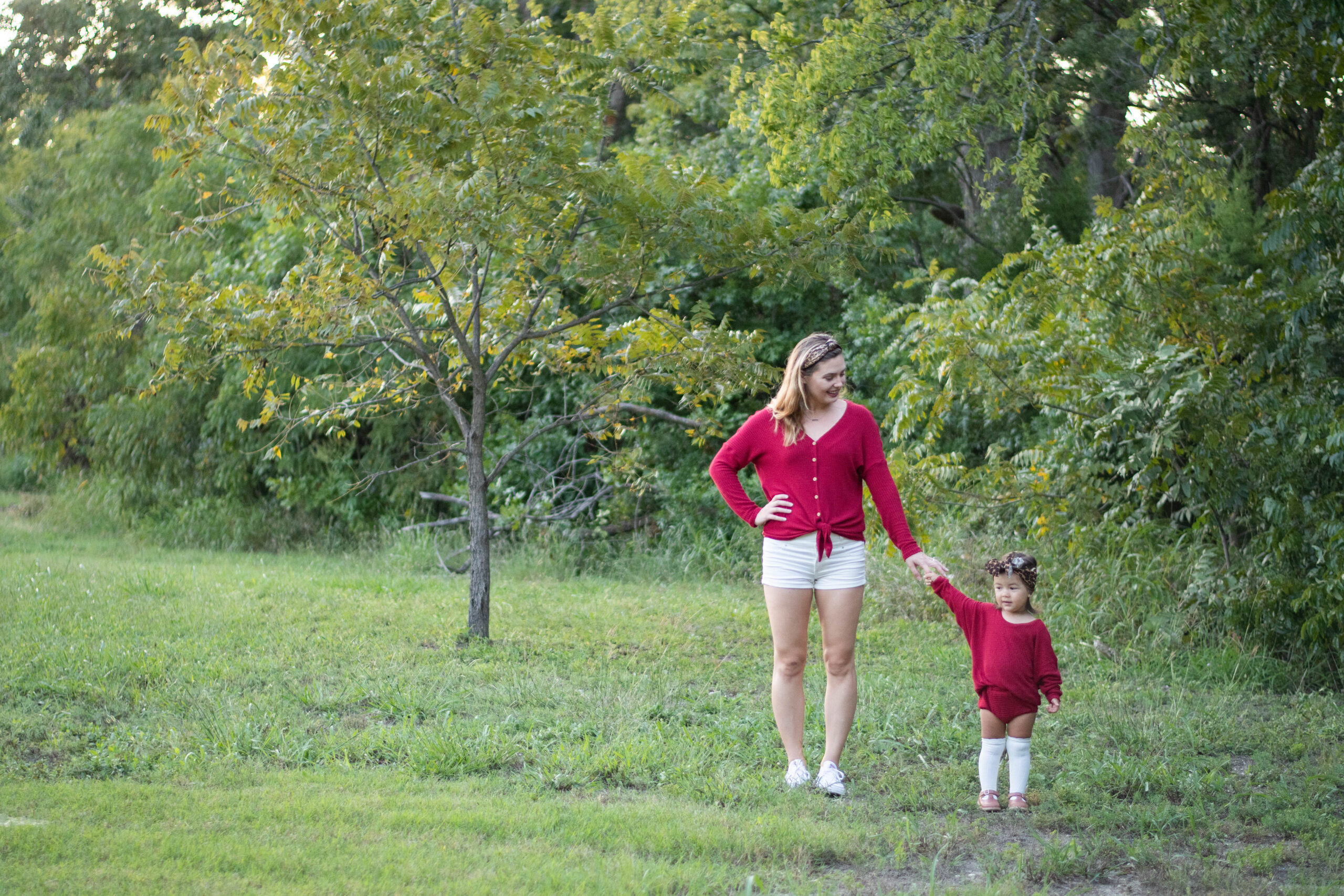 Elizabeth and her daughter stand in a field in front of some green trees with just a hint of autumn coloring on the tips of their leaves. Elizabeth wears a top that she has sewn, and her daughter wears a matching outfit, sewn by Mama, as they hold hands.